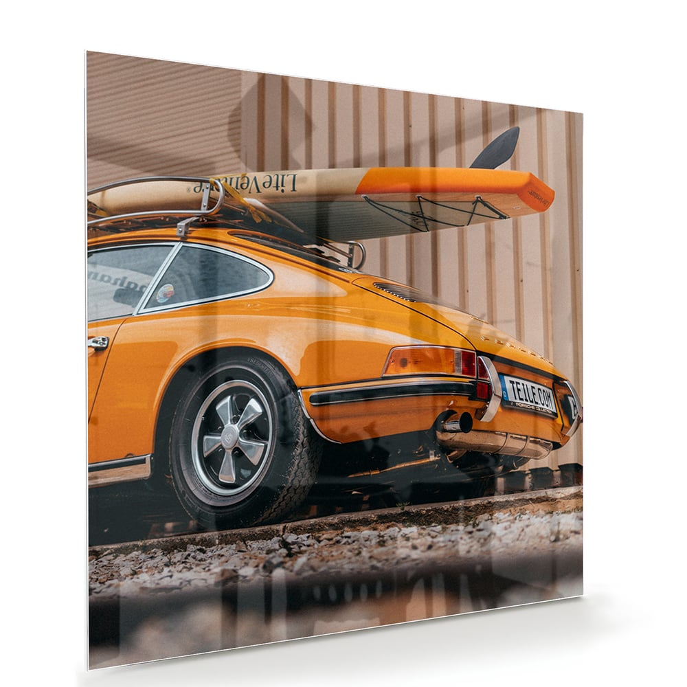 Een glazen schilderij van een oranje auto met een surfbord op het dak, gepresenteerd in een moderne woonkamer.