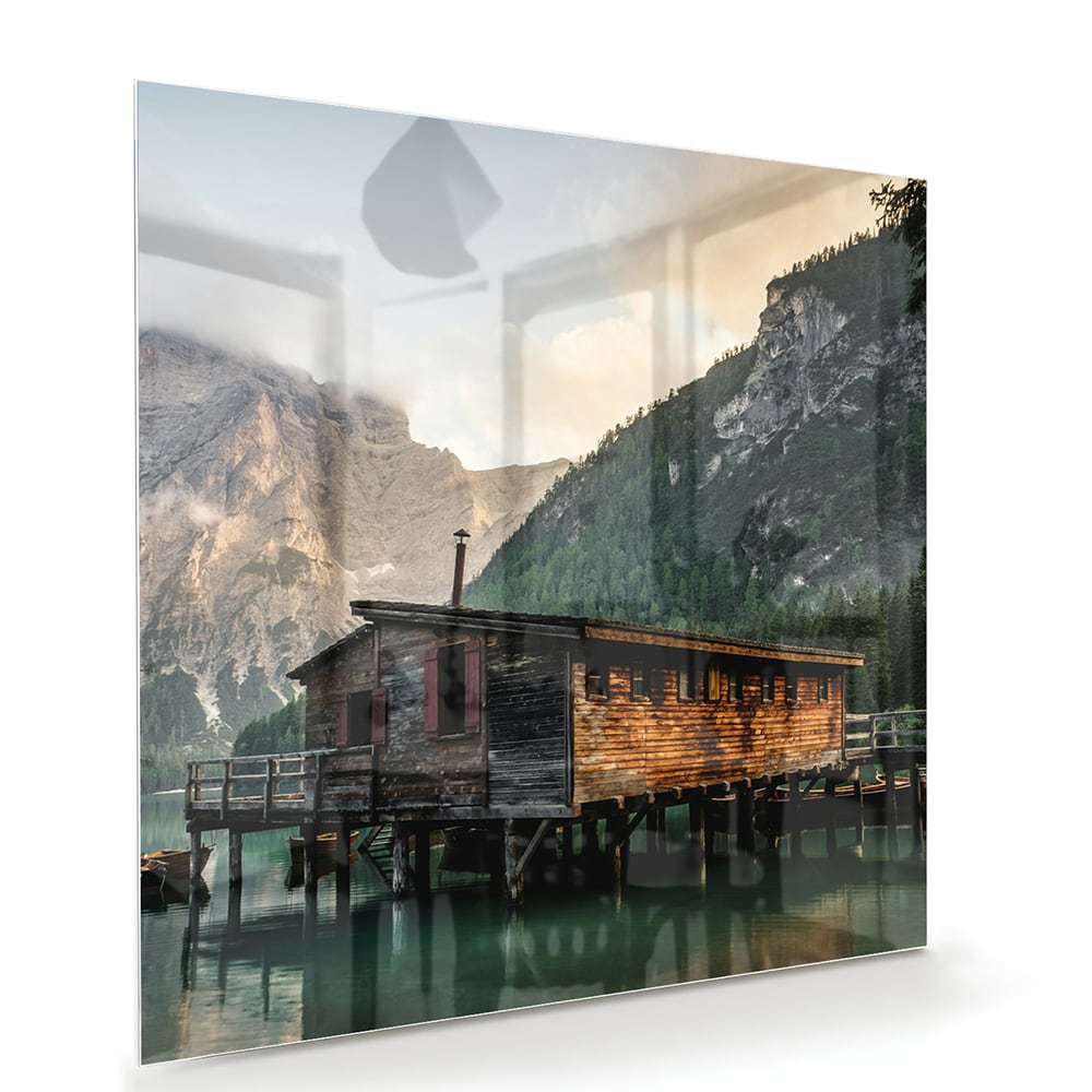 Glazen schilderij van een berghut aan een kalm meer met bergen op de achtergrond.