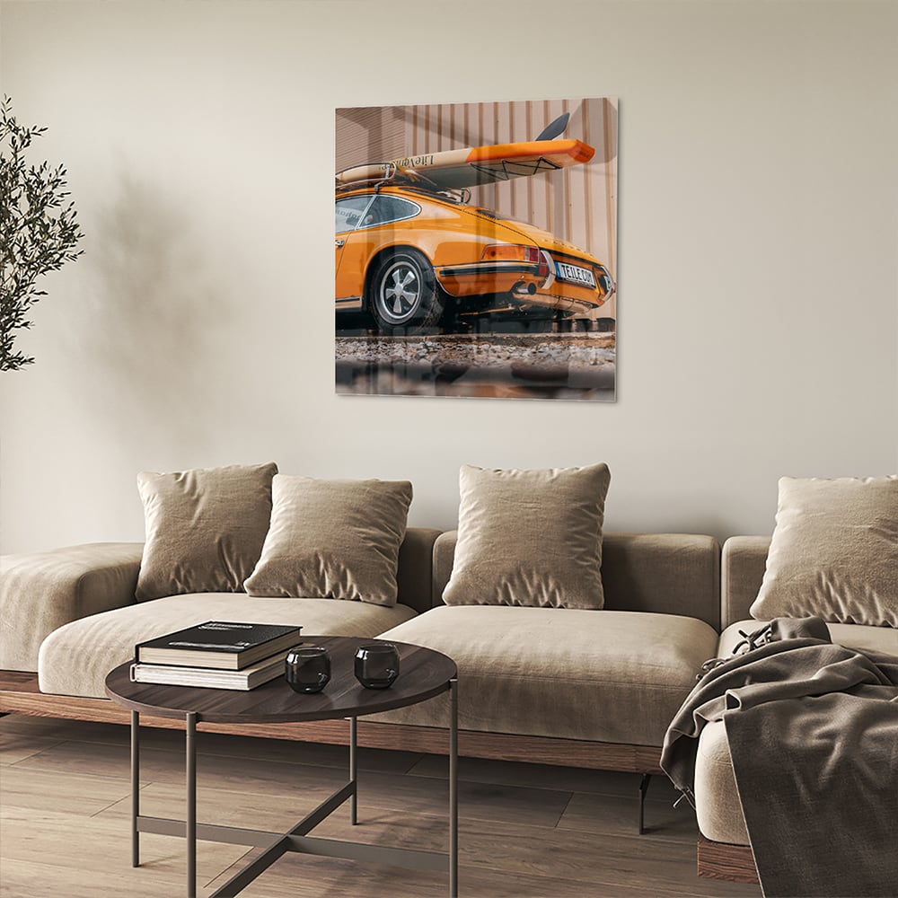 Een glazen schilderij van een oranje auto met een surfbord op het dak, gepresenteerd in een moderne woonkamer.