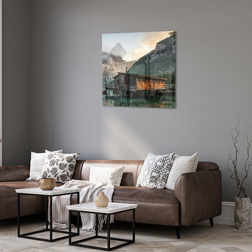 Glazen schilderij van een berghut aan een kalm meer met bergen op de achtergrond.