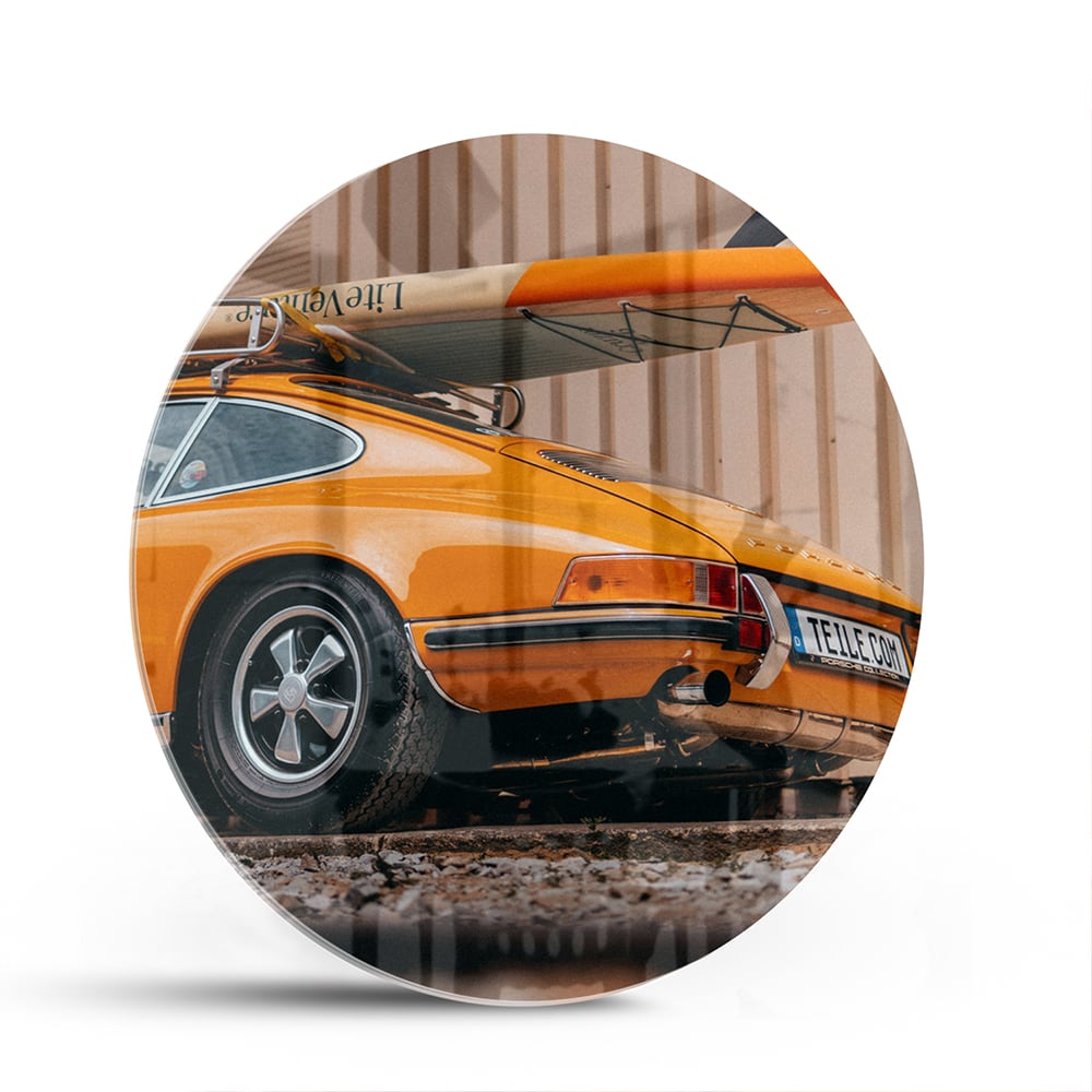 Een glazen schilderij van een oranje auto met een surfbord op het dak, gepresenteerd in een moderne woonkamer.