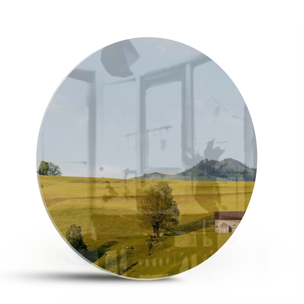 Glazen schilderij met groene heuvels en boerderijen op een witte muur.